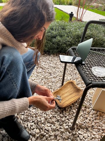 A woman with long brown hair, wearing a beige sweater and blue jeans, sits outdoors on a stone-covered patio. She is focused on assembling or examining Italian charm bracelet links, holding one in her hands above a wooden tray filled with small silver charms. A black smartphone with a green stand rests on a woven chair beside her, and the background shows neatly trimmed greenery and a garden path.