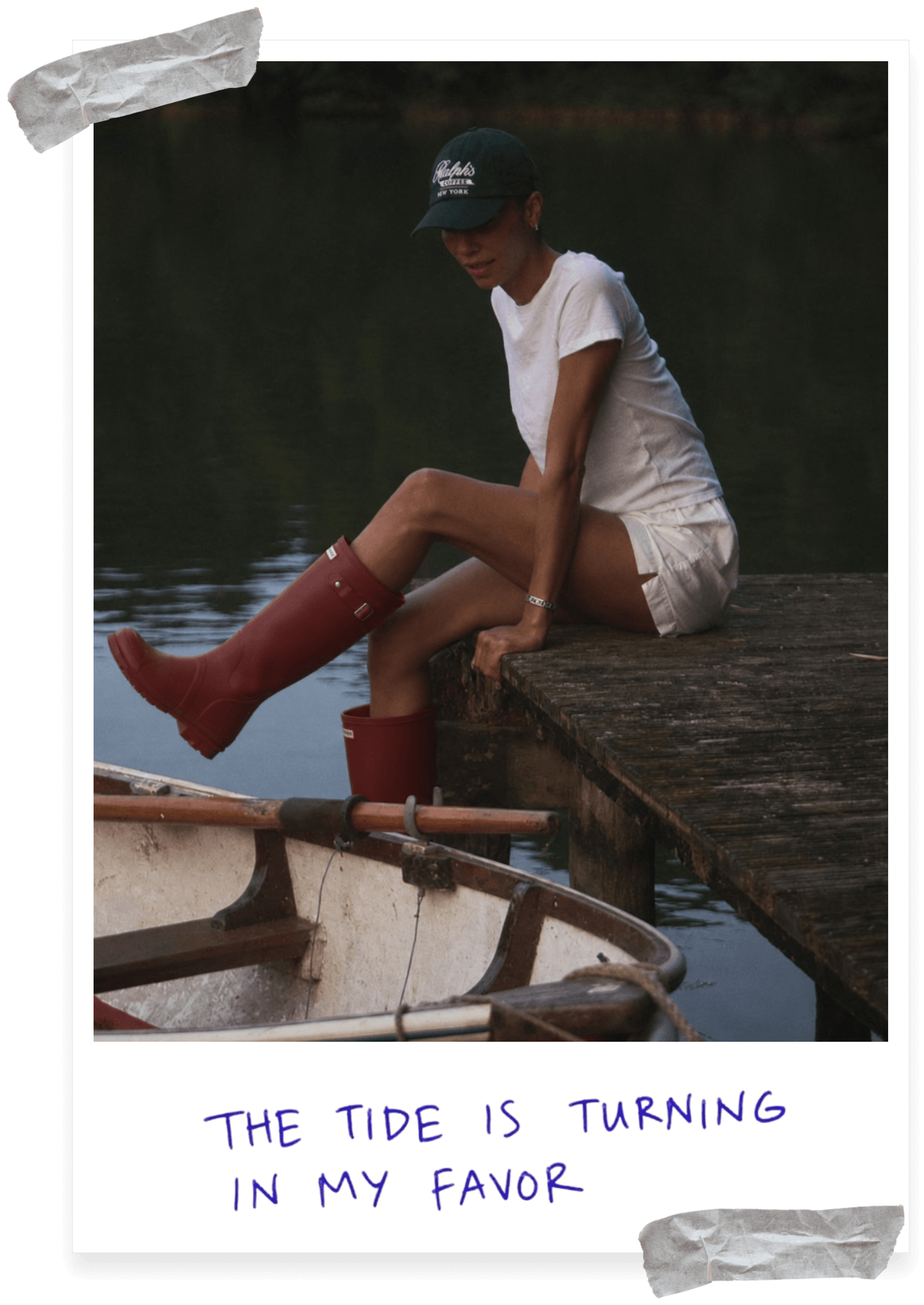 Polaroid-style photo of a person in a green cap, white t-shirt, and white shorts sitting on a wooden dock at dusk, with one leg extended in a red rain boot over a small rowboat. Handwritten caption reads "The tide is turning in my favor."