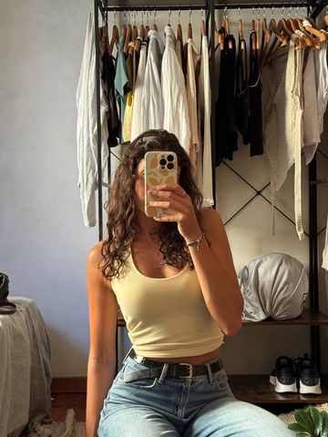 A woman with curly brown hair taking a mirror selfie in a bedroom closet, wearing a yellow crop tank top, wide-leg jeans with a black belt, and a Chericherilady italian charm bracelet visible on her wrist, with a clothing rack full of garments behind her.