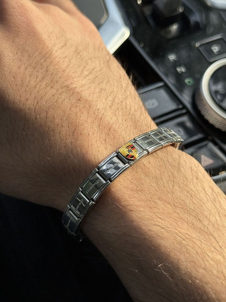A man's wrist wearing a Italian charm bracelet with mostly plain silver links and a single Porsche charm in gold, red, and black, photographed inside a luxury car with a gear console visible in the background.