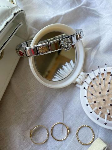 A silver Chericherilady Italian charm bracelet with enamel charms including a camera and a flower, resting on top of a small round compact mirror, laid on a white fabric surface alongside three delicate gold rings and a white mini hairbrush.