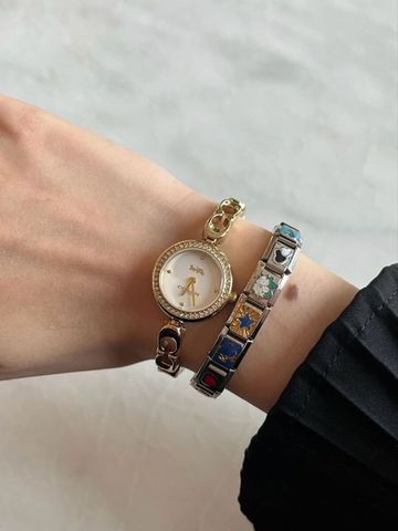 A close-up of a wrist wearing a dainty gold Coach watch with a crystal-encrusted bezel and pearl dial, paired with a silver Chericherilady Italian charm bracelet featuring colorful enamel charms including stars, hearts, and flowers, over a black sleeve against a marble background.