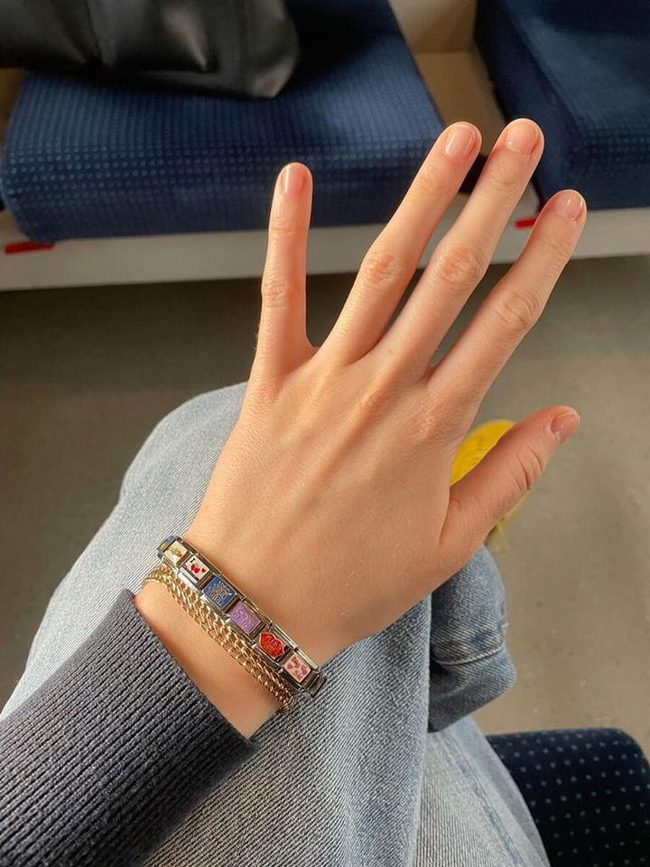 A woman's hand raised to show a stacked bracelet combination on her wrist, featuring a silver Chericherilady Italian charm bracelet with colorful charms and a layered gold chain bracelet, worn over a grey knit sleeve with jeans, seated on public transport with blue upholstered seats in the background.