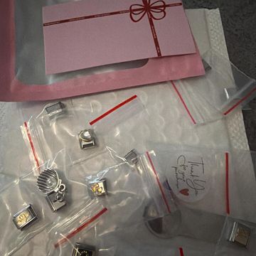 Flat lay of Italian charm bracelet pieces packaged in small clear zip bags on top of soft white padding. A pink thank-you card with a red ribbon bow design sits above the charms