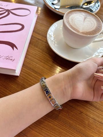 A woman's wrist wearing a silver Chericherilady Italian charm bracelet with colorful charms, resting on a wooden table beside a latte with heart latte art in a white cup and saucer, and a pink hardcover notebook with a bow design.