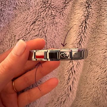 Hand holding a silver Italian charm bracelet, featuring a poland flag charm, a black charm, and a silver heart charm, against a soft textured fabric background.