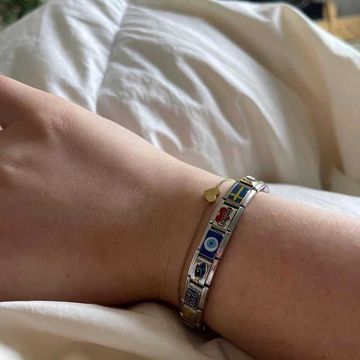 Silver Italian charm bracelet with colorful links including Sweden flag, evil eye, Coca-Cola logo, blue dolphin, and I love Dad charm, worn next to a delicate gold bracelet with a small heart charm