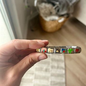 Women hand holding a silver Italian charm bracelet with colorful enamel charms including a cactus, truck, face, flag, four-leaf clover, and red heart shown in a soft interior background.