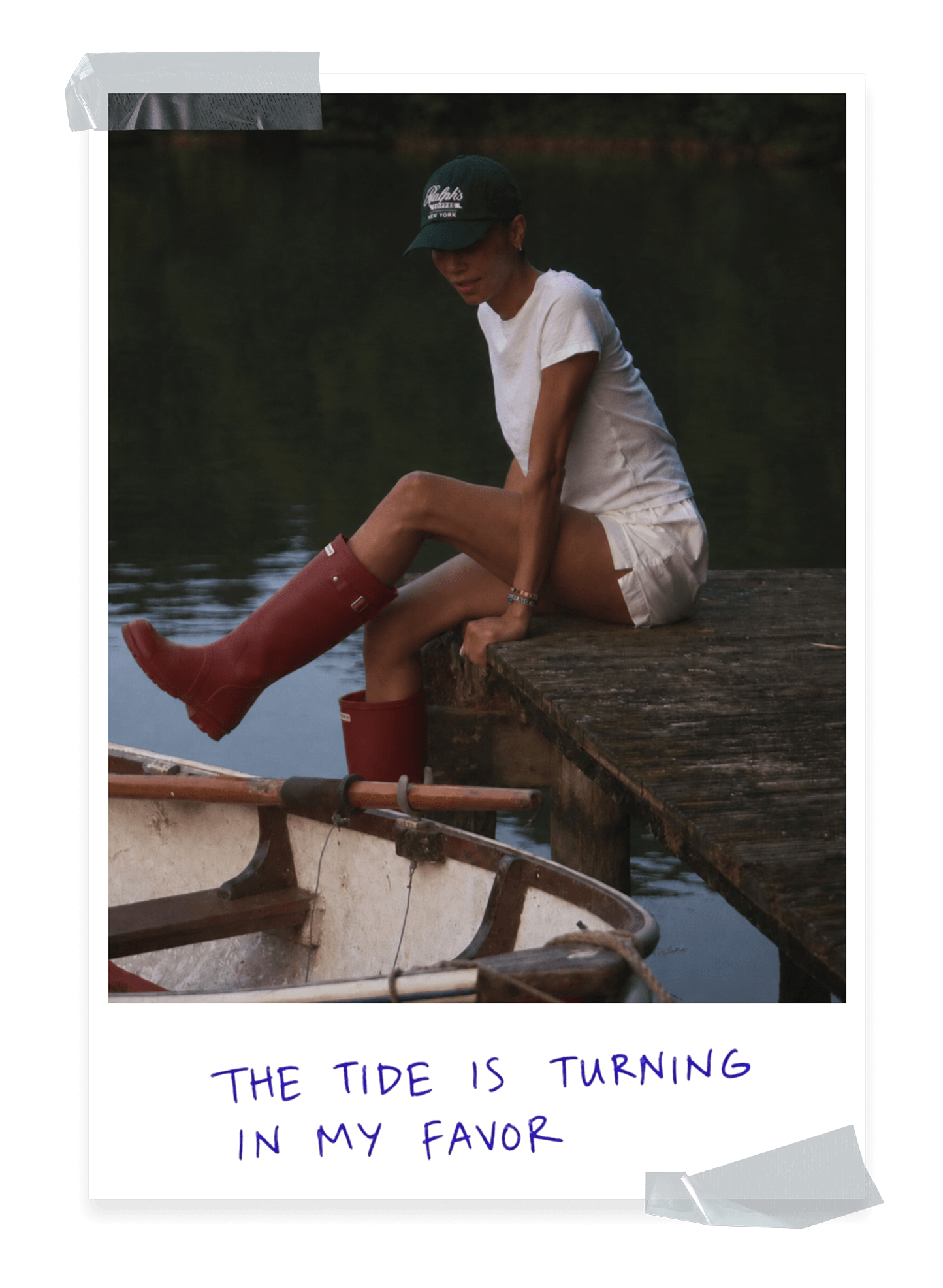 Polaroid-style photo of a person in a green cap, white t-shirt, and white shorts sitting on a wooden dock at dusk, with one leg extended in a red rain boot over a small rowboat. Handwritten caption reads "The tide is turning in my favor."